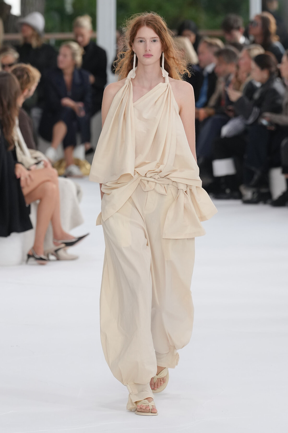 Model walks the runway in a cream draped halter top with matching wide, gathered pants and beige sandals.
