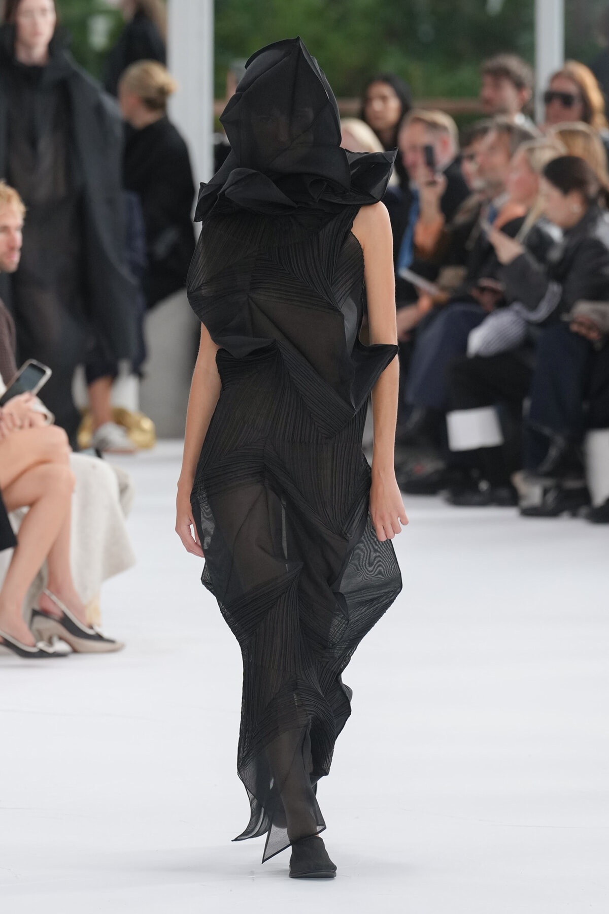Model walking a runway in a black sculptural gown with pleated geometric panels and an oversized hood; audience in the background.
