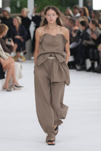 Model wearing taupe draped halter top and billowy wide-leg pants on a fashion runway with a blurred audience behind her.