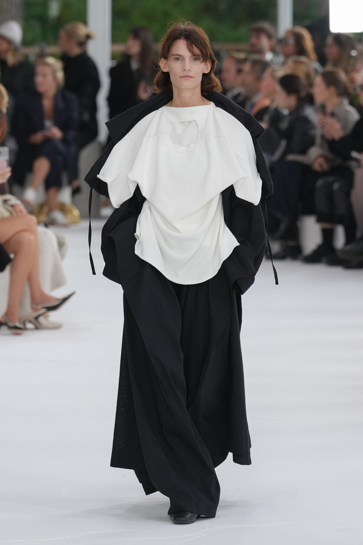 Model walking a fashion show runway in an oversized white top layered over black wide pants; audience seated in background