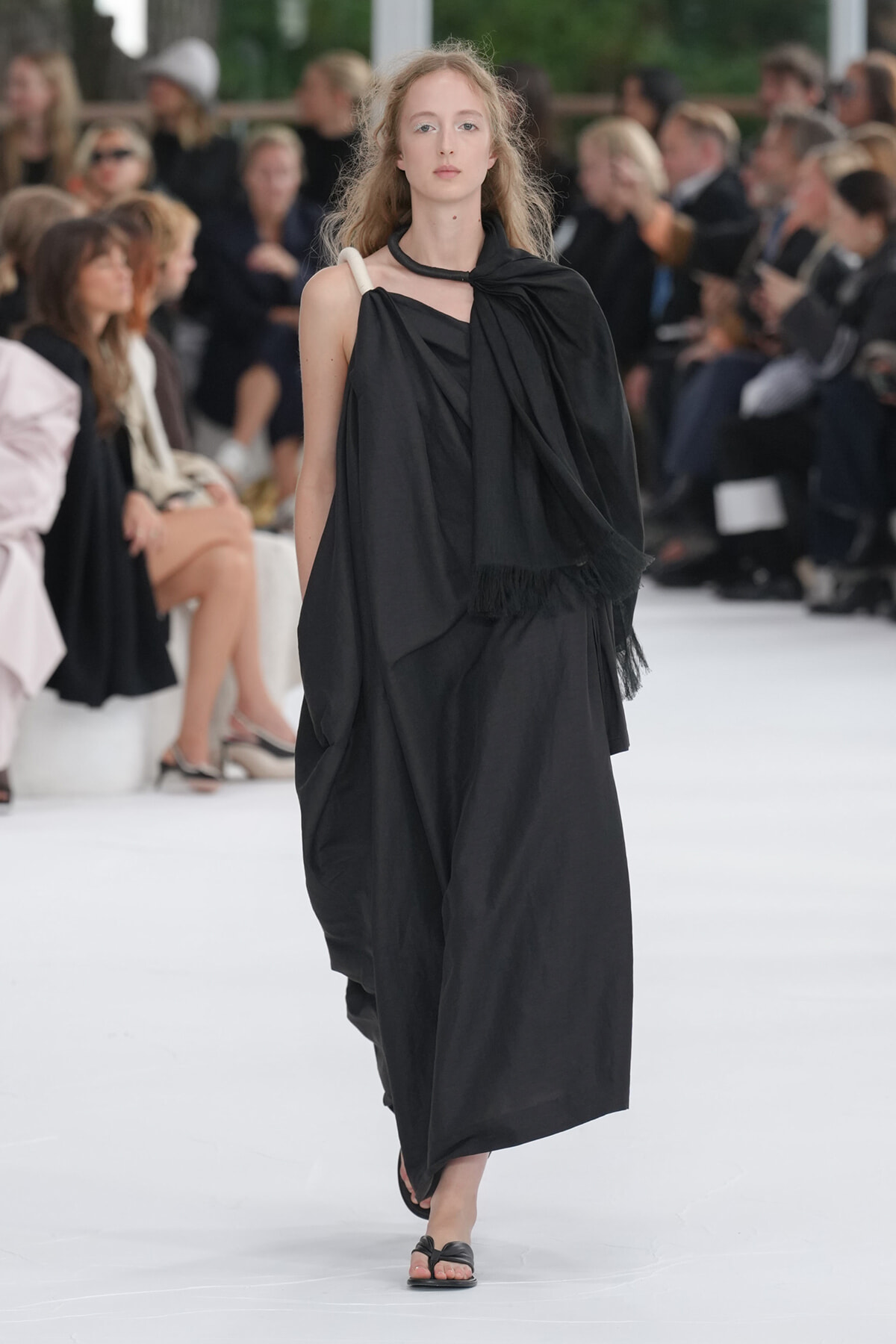 Model walks a fashion runway in a loose, black draped dress with a scarf-like wrap and flat sandals, audience blurred in the background.