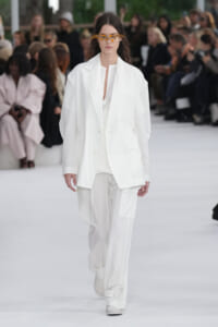 Model in an oversized white suit walks a runway, with an orange flower headpiece across her eyes.