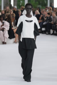 Dark-skinned model on a runway wearing a white short-sleeve top over a black oversized layer and wide black pants, with sunglasses; blurred audience in background.