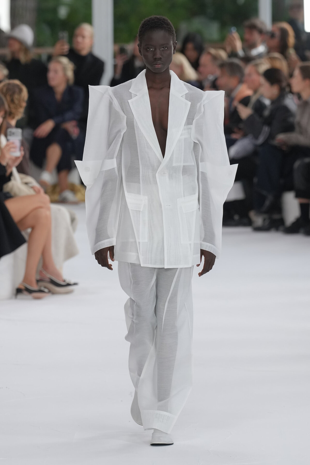 Model walking a fashion show in an oversized white blazer with broad, structured shoulders and sheer pants, audience in the background.