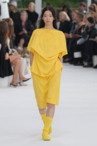 Model walks the runway in a bright yellow, draped top with a circular neckline and matching knee-length shorts, yellow socks visible; audience blurred in background.