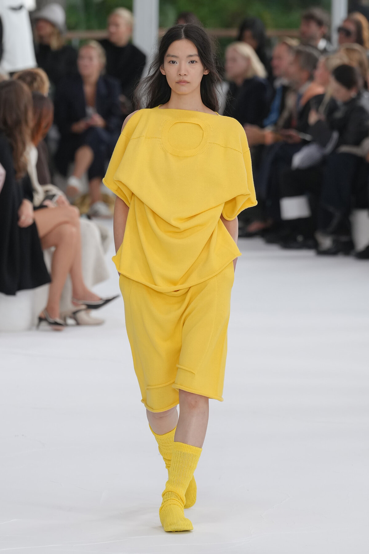 Model walks the runway in a bright yellow, draped top with a circular neckline and matching knee-length shorts, yellow socks visible; audience blurred in background.