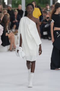 Model walks a runway in a white one‑shoulder dress, carrying a folded white jacket and wearing white knit socks.