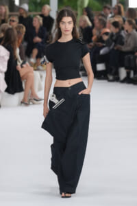 Model walking the runway in a black cropped top and oversized black cargo pants with a white tool accessory clipped at the hip, audience in background.