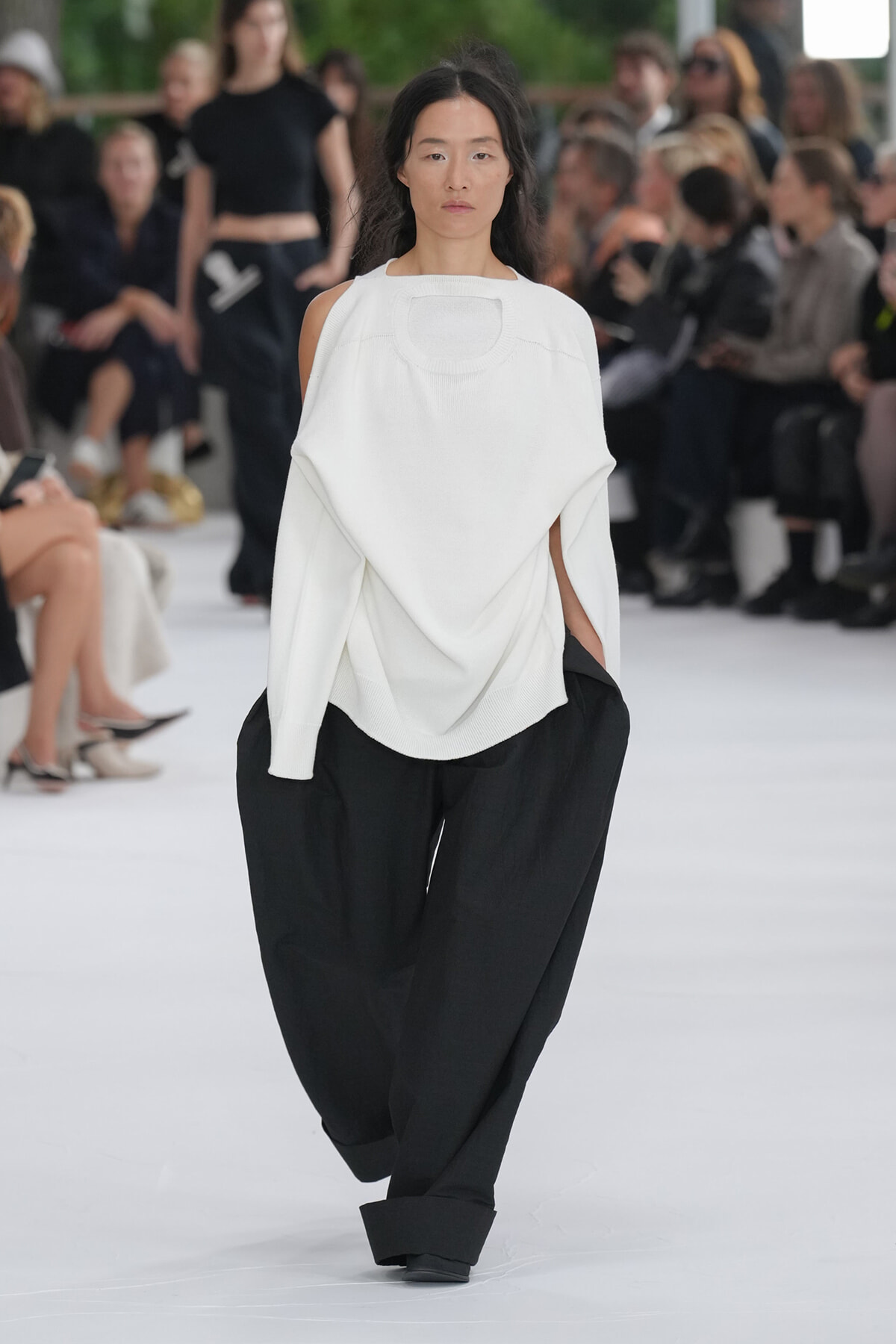 Model walking a fashion runway in a loose white poncho-style sweater and wide black pants, with seated audience in background.