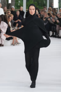 Female model walks a runway in an all-black, sculptural pleated outfit with cape-like sleeves, extending her left arm to showcase the draped fabric.