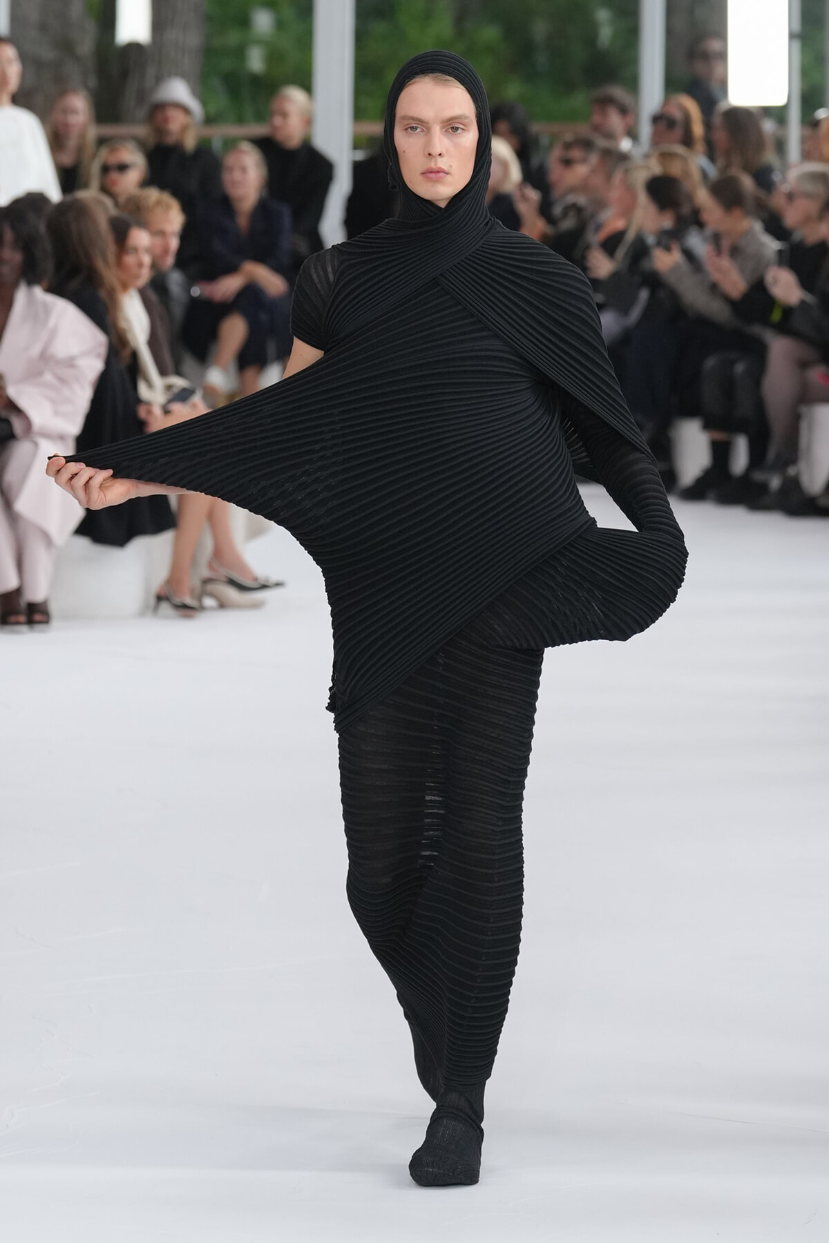 Female model walks a runway in an all-black, sculptural pleated outfit with cape-like sleeves, extending her left arm to showcase the draped fabric.