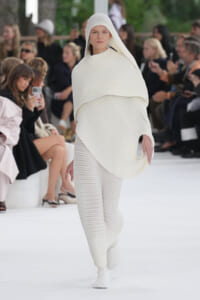 Model walks a fashion runway wearing a white, ribbed knit hooded poncho and matching pants.