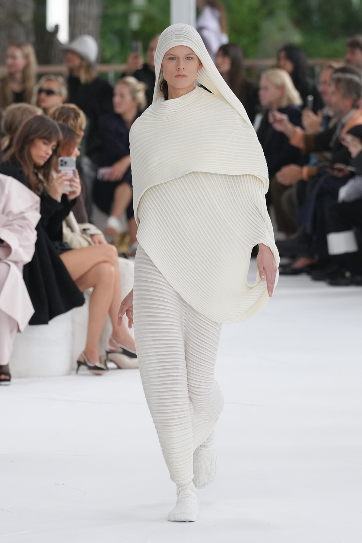 Model walks a fashion runway wearing a white, ribbed knit hooded poncho and matching pants.