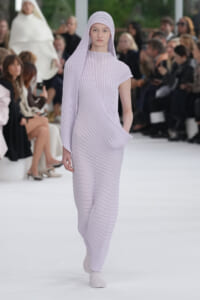 Model wearing lavender ribbed pleated dress with matching headscarf walking a fashion runway, audience visible in background.