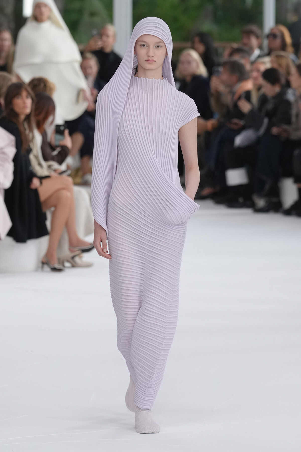 Model wearing lavender ribbed pleated dress with matching headscarf walking a fashion runway, audience visible in background.