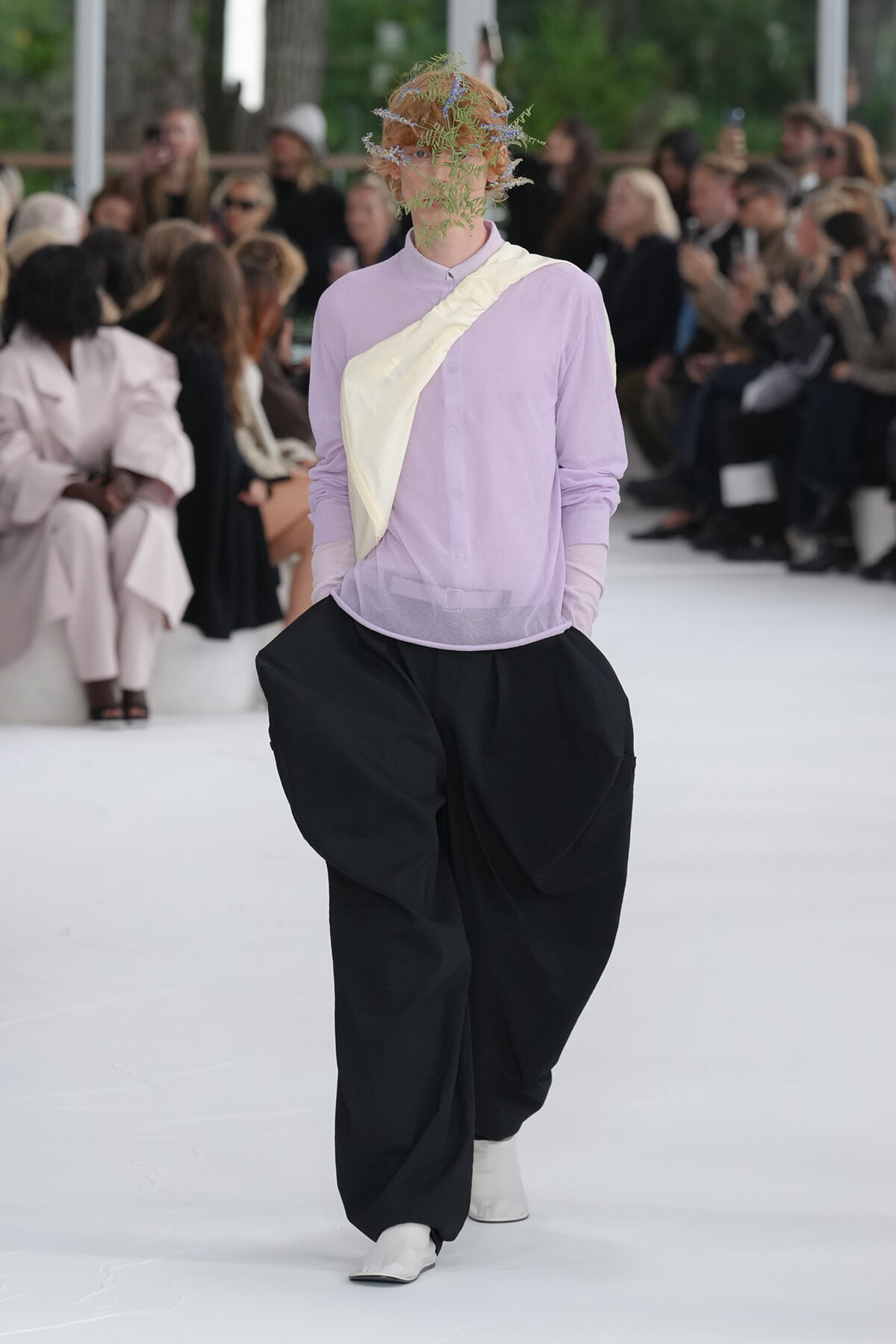 Model walking a runway in a lavender button-up with a pale yellow diagonal sash, oversized black pants, white boots, and a botanical headpiece. Audience blurred in background.