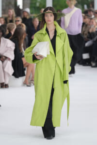 Model walks runway in a neon lime-green long trench over a black outfit, holding a white clutch with a leafy hat adorned with greenery above her head.