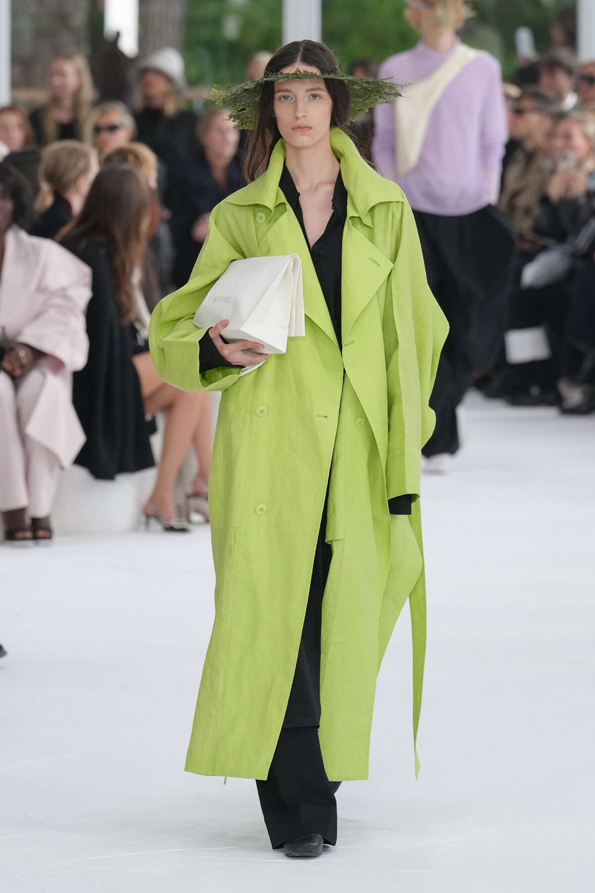 Model walks runway in a neon lime-green long trench over a black outfit, holding a white clutch with a leafy hat adorned with greenery above her head.