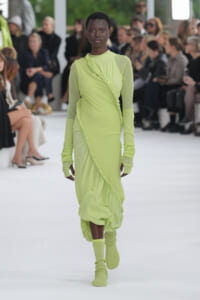 Model walking a white runway in a neon lime-green dress with rope detailing and matching knit sleeves and socks; audience in the background.