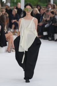 Model walks a runway in an oversized white top and wide black pants, with a leafy flower mask covering her eyes for a fashion show touch.