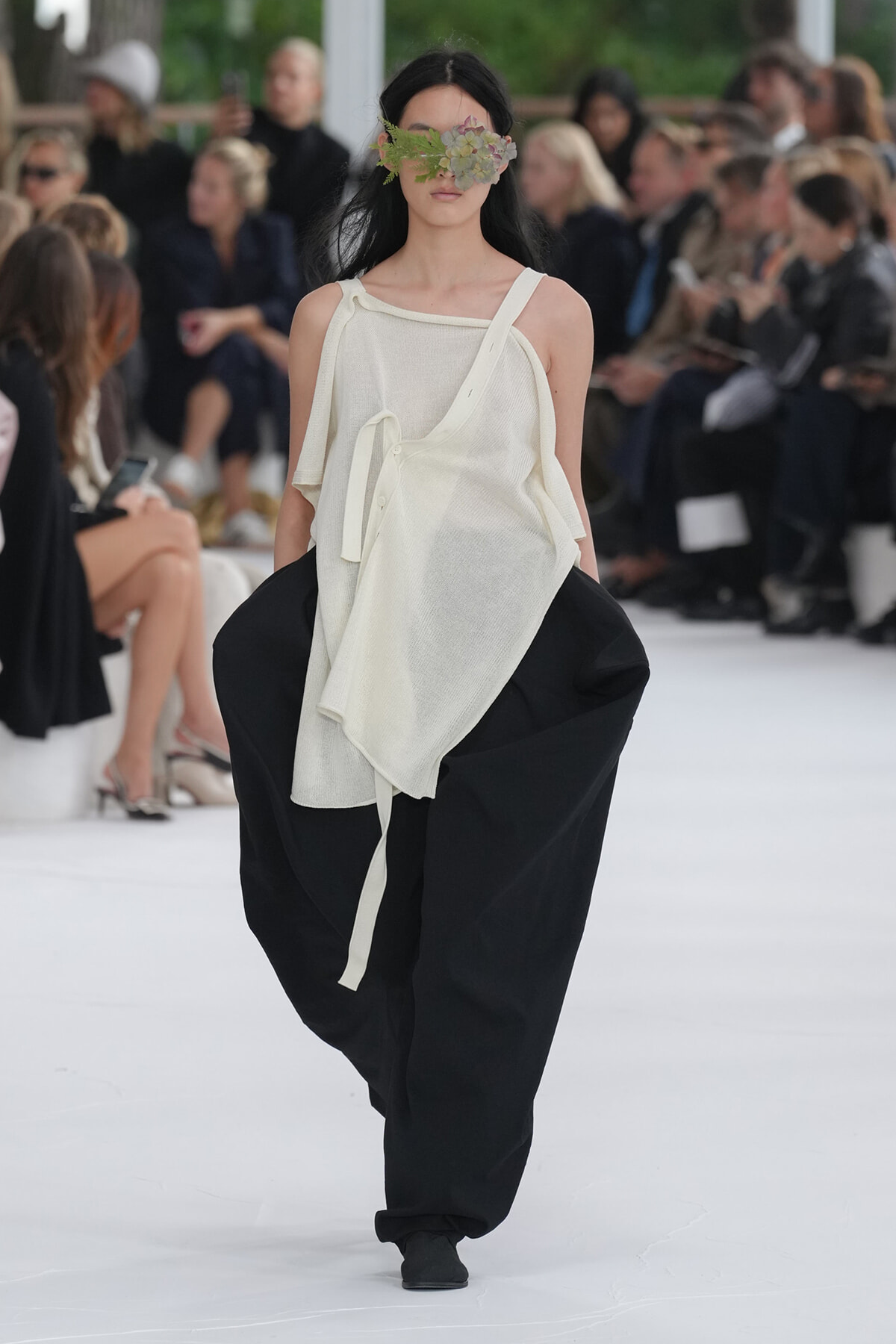Model walks a runway in an oversized white top and wide black pants, with a leafy flower mask covering her eyes for a fashion show touch.