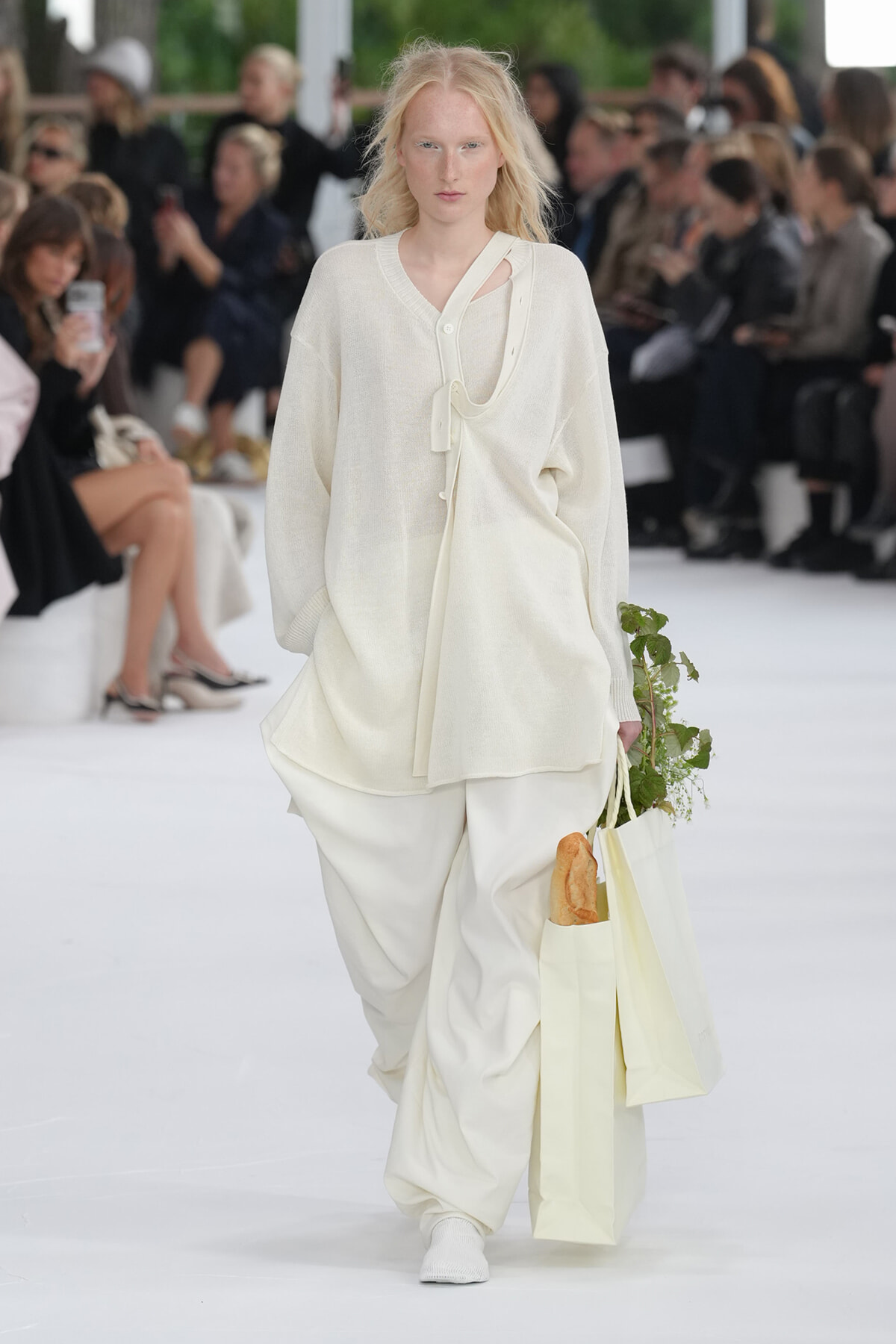 Model walking a white runway in an all-white knit ensemble; carrying a cream tote with a baguette and greens peeking out.