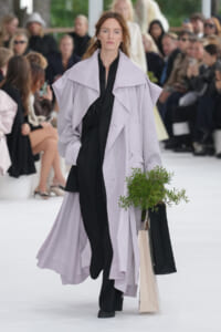 Model walks a runway in a pale lavender oversized coat over a black outfit, holding a potted plant in a beige shopping bag.
