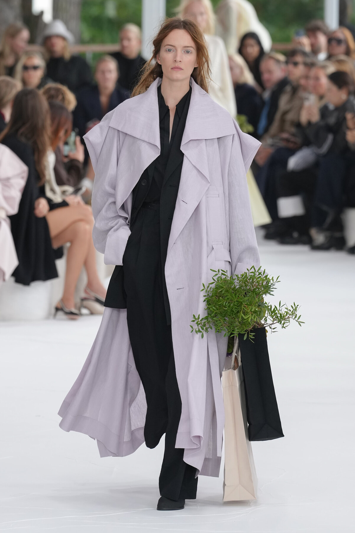 Model walks a runway in a pale lavender oversized coat over a black outfit, holding a potted plant in a beige shopping bag.