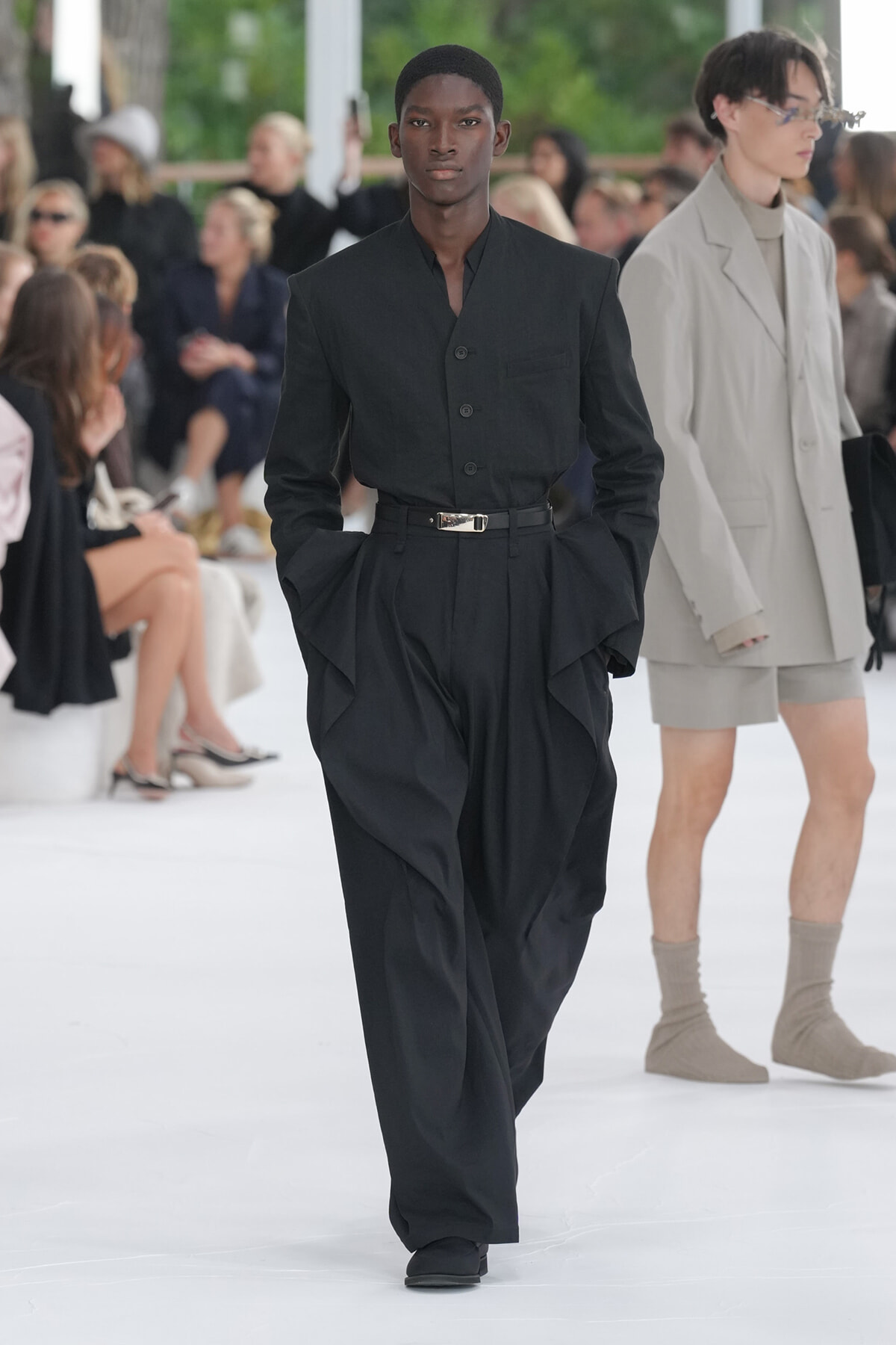 Model walking the runway in a black tailored shirt and wide-legged pants with a silver belt; audience blurred in background.