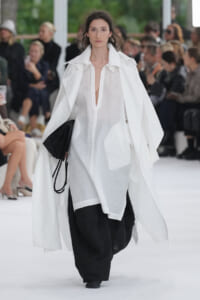 Model on a runway wearing a sheer white blouse with a flowing white cape over wide black pants; audience blurred in background.