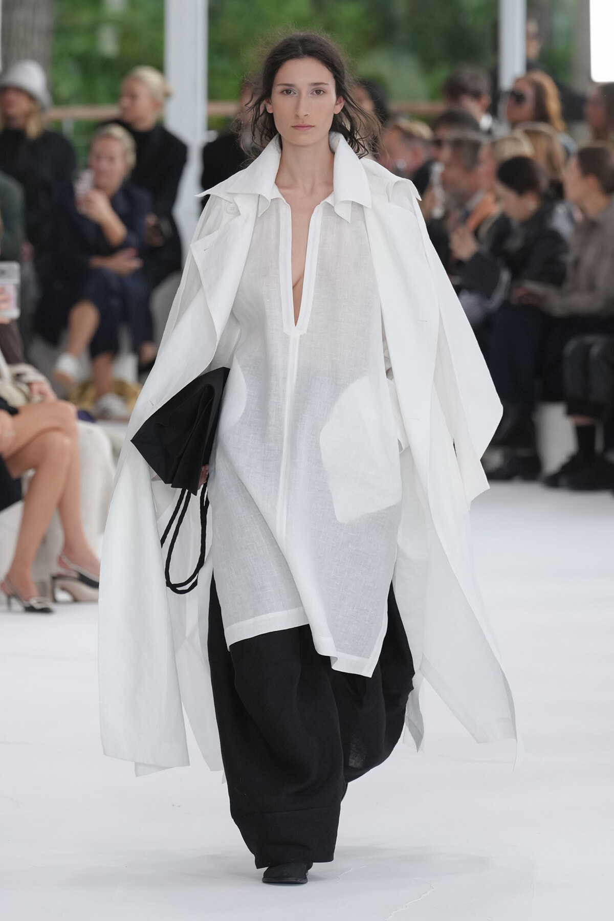 Model on a runway wearing a sheer white blouse with a flowing white cape over wide black pants; audience blurred in background.