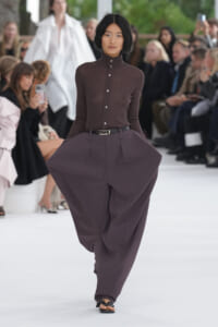 Model walks the runway in a fitted brown high-collar button-up top with oversized purple-brown wide-leg pants and a black belt.