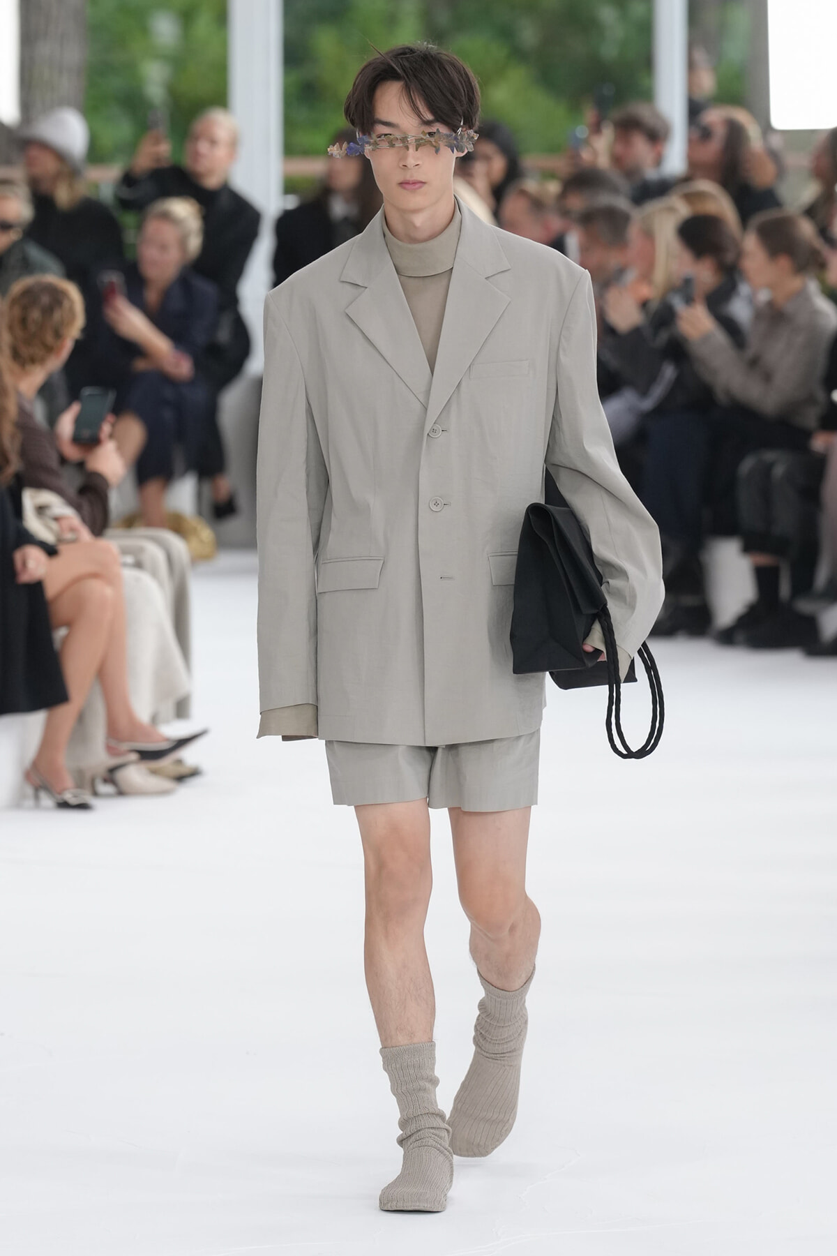 Model walking the runway in a light gray oversized blazer and matching shorts, beige turtleneck, flower-embellished eyewear, carrying a black bag; audience in background.