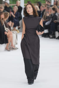Model walking a fashion runway in a dark sleeveless dress with draped detailing, holding a black clutch; audience blurred in the background
