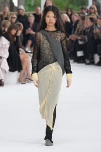 Model walking runway in sheer black lace top with pale yellow draped skirt and gray sash; audience blurred in background.