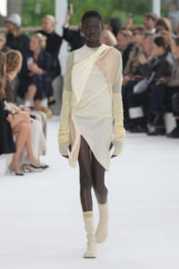 Black model walks a fashion runway in a cream draped dress with pale yellow knit sleeves and matching socks.