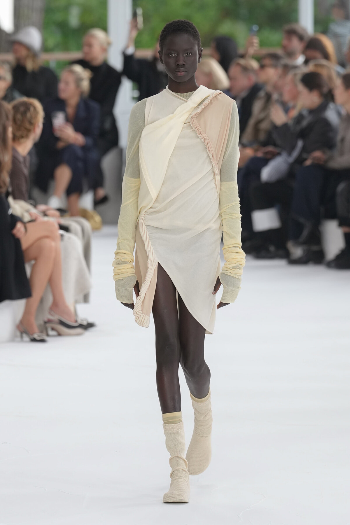Black model walks a fashion runway in a cream draped dress with pale yellow knit sleeves and matching socks.