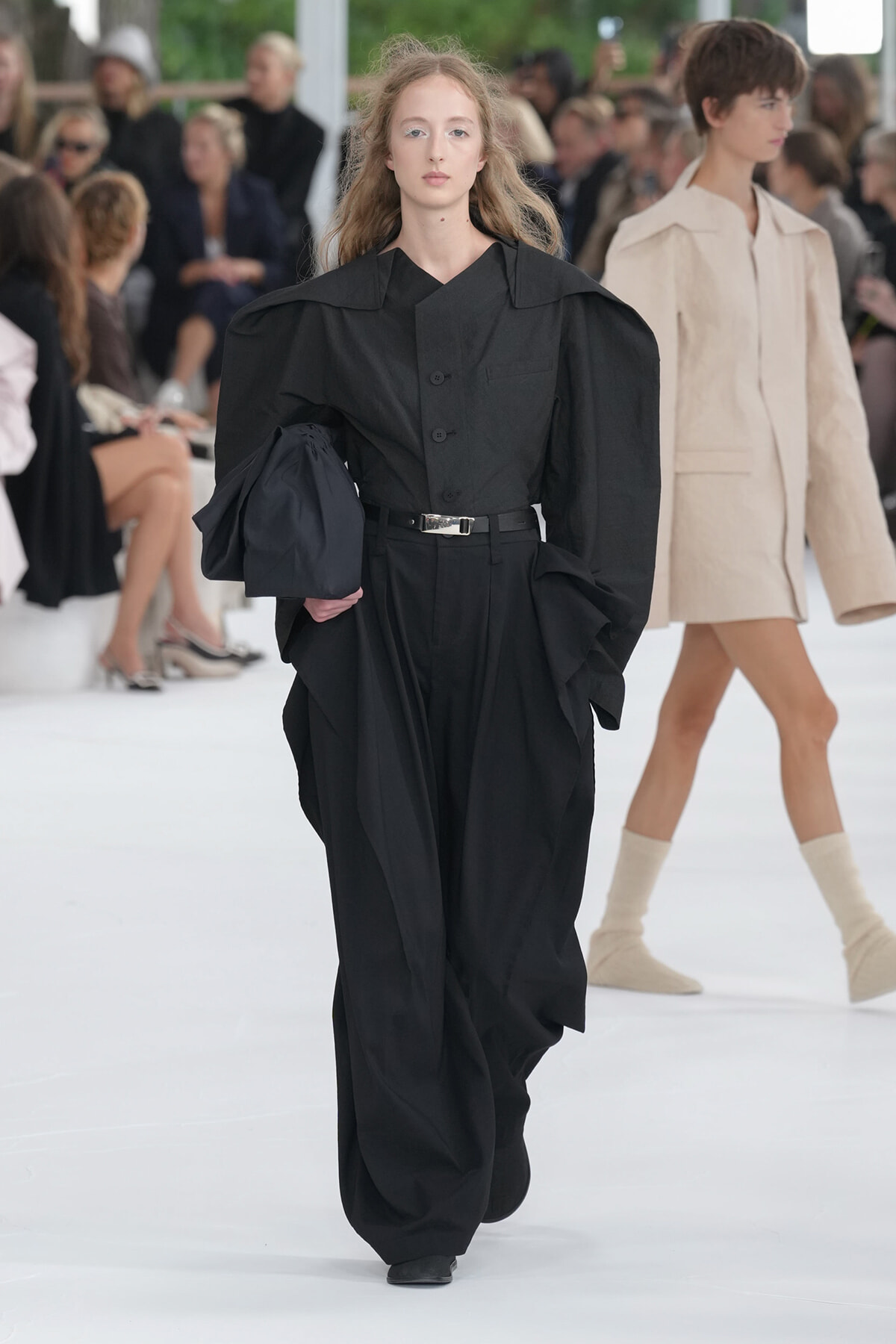 Model walks the runway in an oversized black jumpsuit with dramatic shoulder volume, belted waist, and a dark clutch left arm.