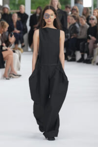 Model walks the runway in a black sleeveless jumpsuit with a belt and oversized, tapered pants; audience blurred in background.