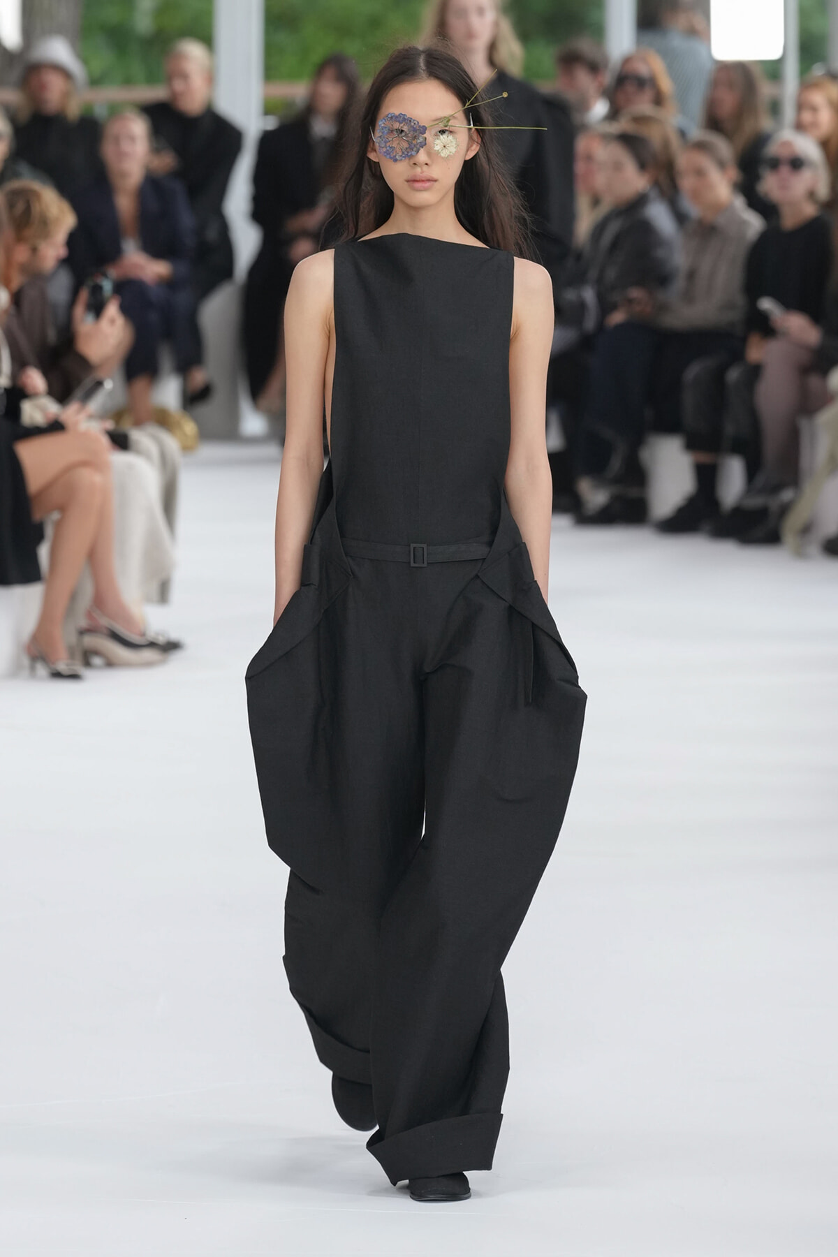 Model walks the runway in a black sleeveless jumpsuit with a belt and oversized, tapered pants; audience blurred in background.