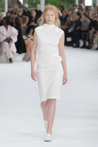 Model walks down a white runway in an all-white sleeveless dress with a high neckline; audience seated along the sides in soft focus.