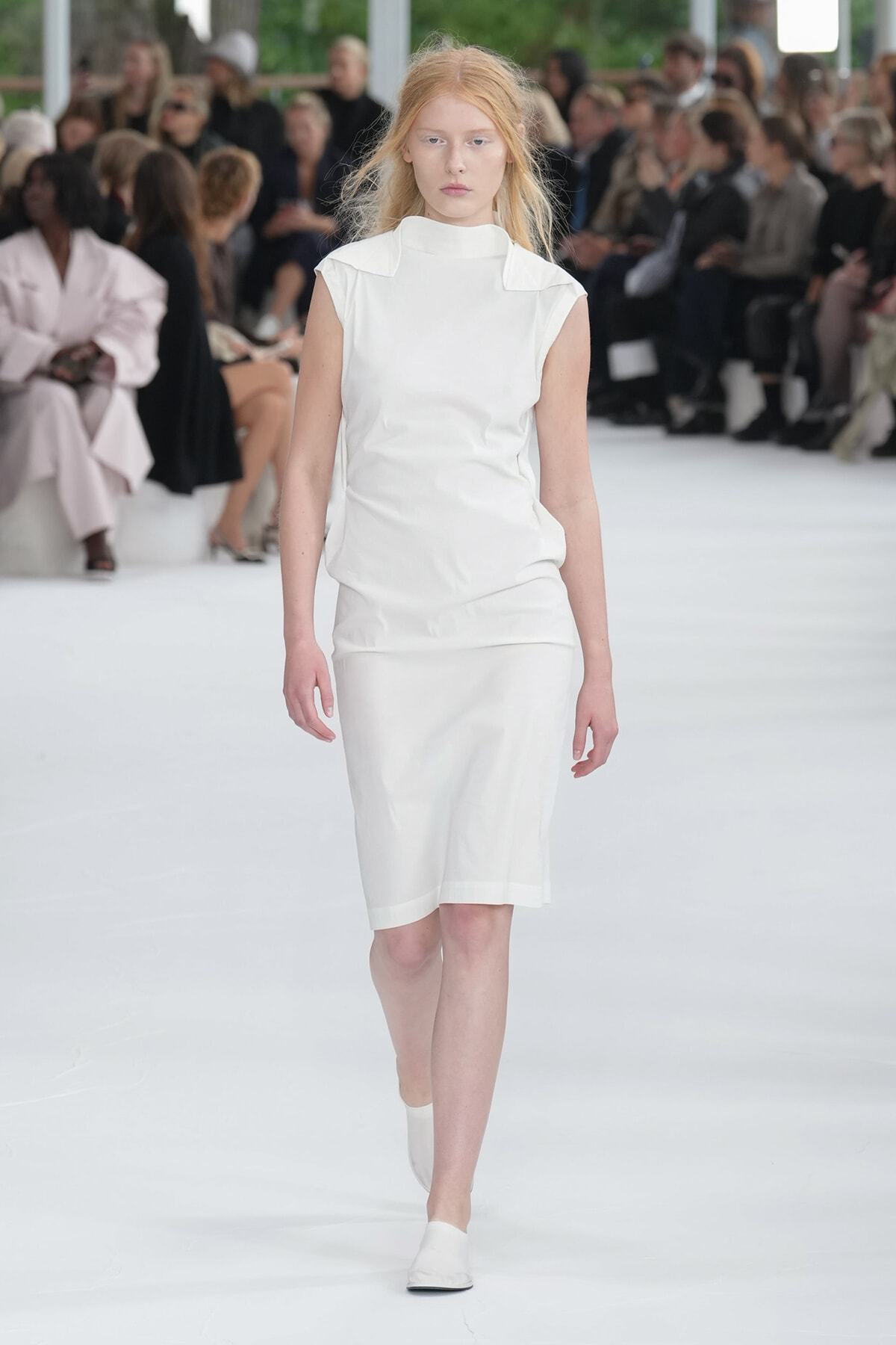 Model walks down a white runway in an all-white sleeveless dress with a high neckline; audience seated along the sides in soft focus.