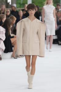 Model walks a fashion runway wearing a beige oversized structured coat with broad shoulders and wide lapels, paired with cream knit socks; audience blurred in the background.