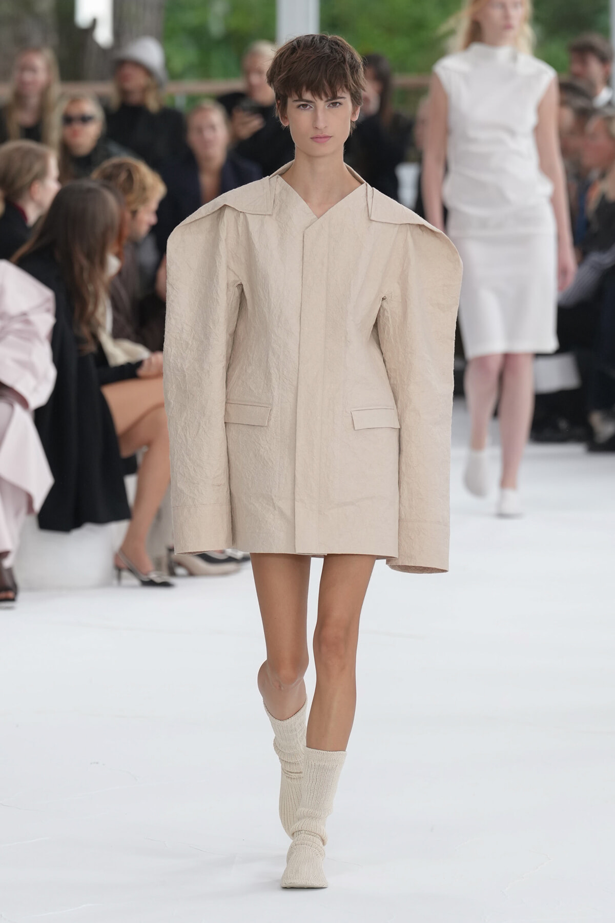 Model walks a fashion runway wearing a beige oversized structured coat with broad shoulders and wide lapels, paired with cream knit socks; audience blurred in the background.