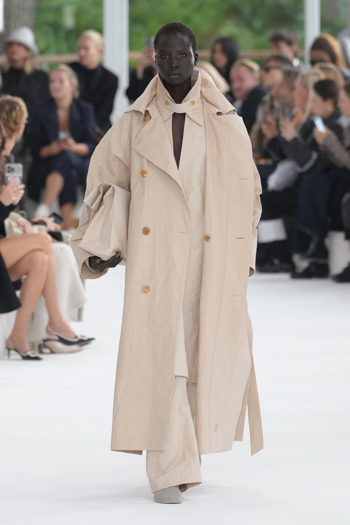 Model walks the runway wearing a large beige trench coat with double-breasted buttons; audience watches in background.