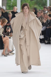 Model walks a fashion runway in an oversized beige trench coat with layered beige outfits, while a blurred audience watches in the background.