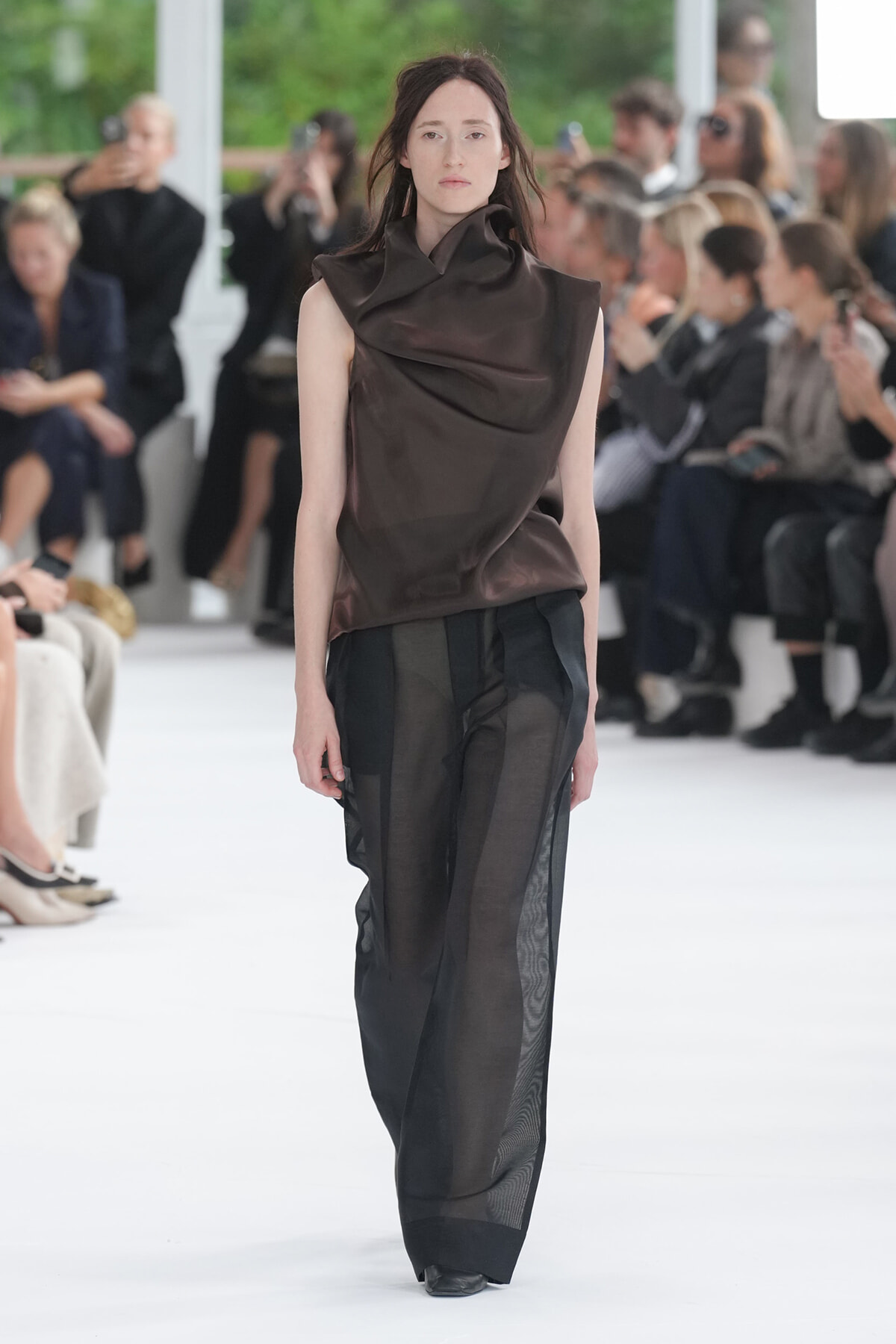 Model walks the runway in a brown draped sleeveless top paired with sheer black wide-leg pants, audience blurred in the background