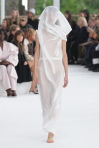Model on a fashion runway wearing a sheer white gown with an oversized veil, walking barefoot.