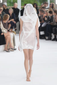 Model walking the runway in a translucent white draped dress with a hooded scarf, audience blurred in the background.