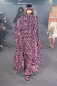 Model walking a runway in a long purple floral dress with a high neck and voluminous sleeves, pink lipstick, and a bold bob hairstyle nearby audience in the background.
